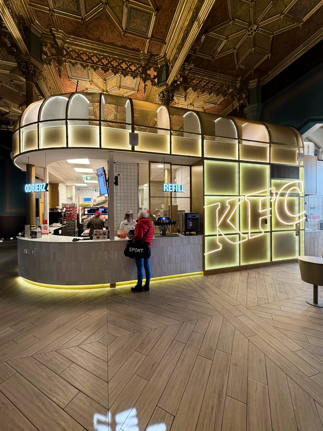 KFC Wrocław Główny — glazed counter pavilion with neon signage under the gilded coffered station ceiling